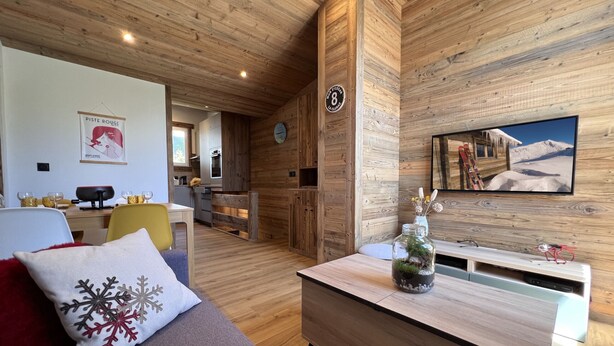 Une pièce chaleureuse avec un plafond en bois, un lit agrémenté d’un oreiller rouge, une petite table sur laquelle se trouve un pot de plantes et un téléviseur diffusant une image d’une montagne enneigée.