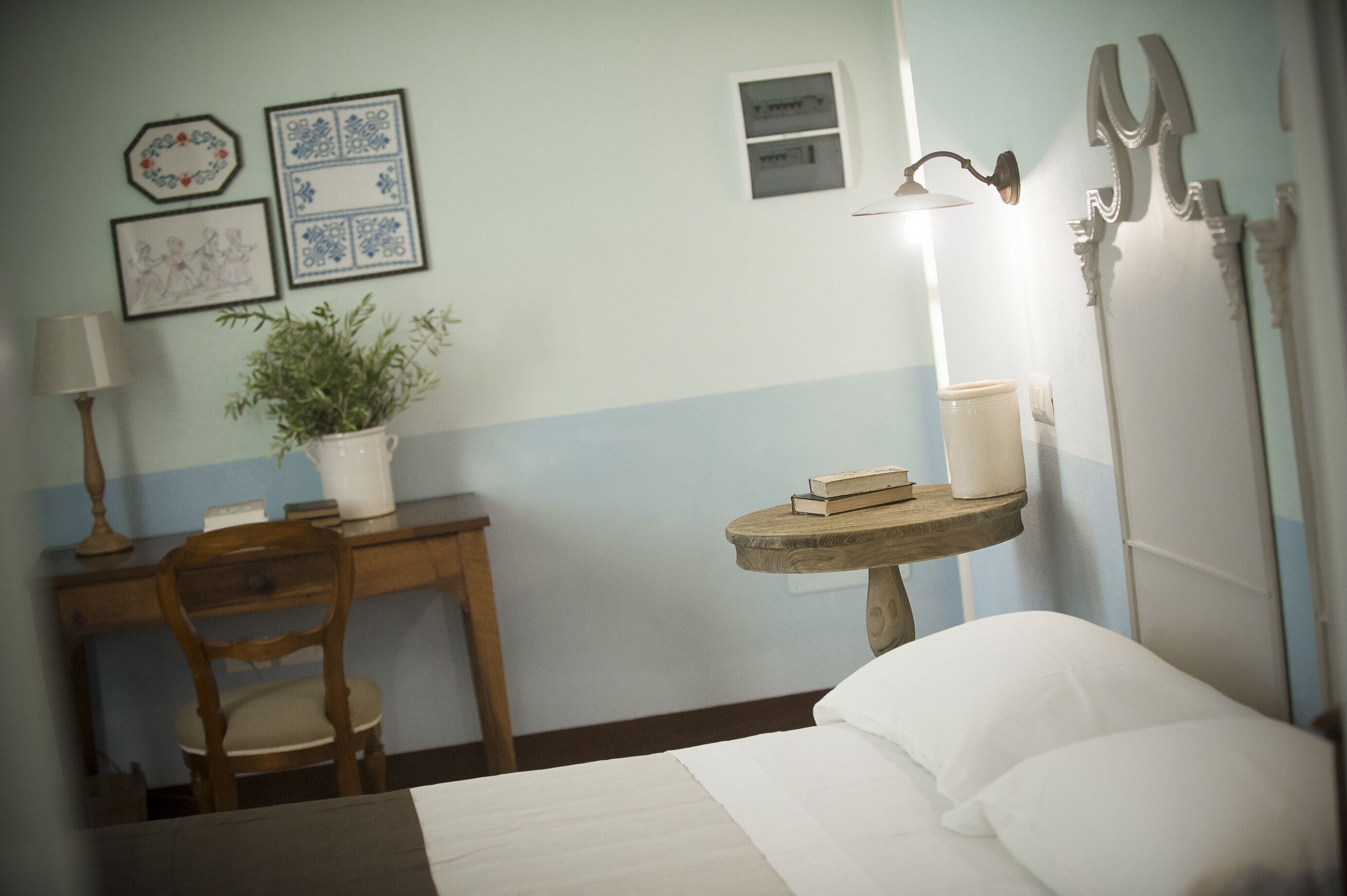 Habitación de hotel con cama, escritorio con lámpara, planta en maceta y mesita de noche con libros y lámpara.