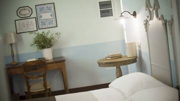 Habitación de hotel con cama, escritorio con lámpara, planta en maceta y mesita de noche con libros y lámpara.