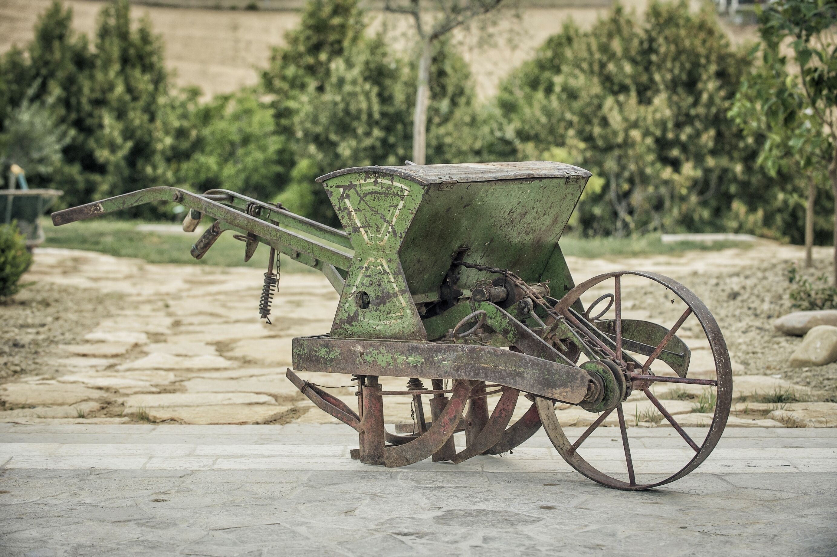 Una máquina agrícola antigua, verde y estacionaria, sobre ruedas, ubicada en una superficie pavimentada en un espacio al aire libre con árboles y un paisaje montañoso al fondo.