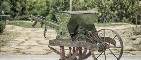 An old, green, stationary agricultural machine with wheels, set on a paved surface in an outdoor area with trees and a hilly background.