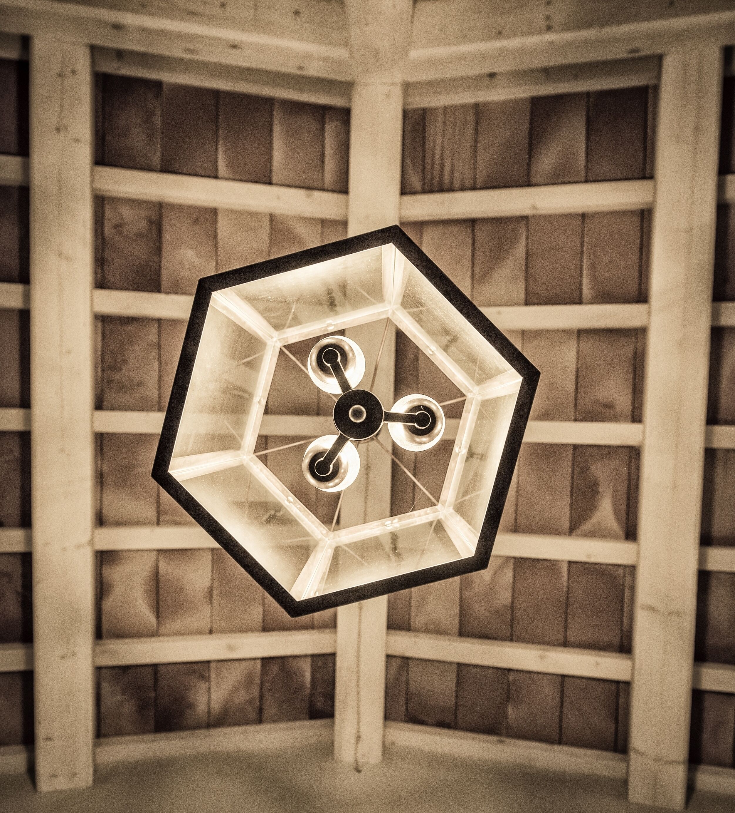 A hexagonal ceiling light fixture with three adjustable arms, set against a wooden wall with a grid pattern.