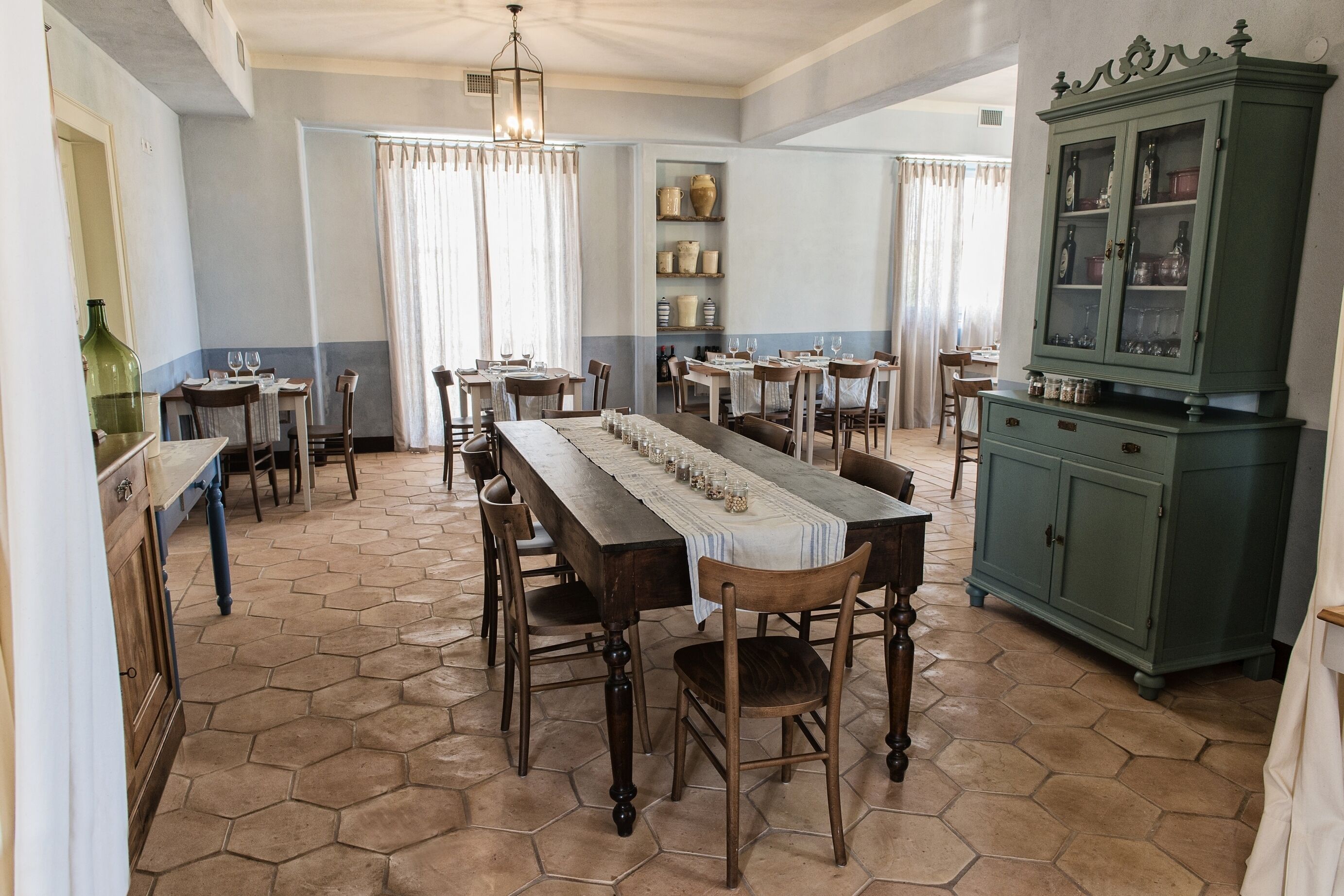 Un comedor con suelo de baldosas hexagonales, mesas de madera con sillas, una mesa larga con mantel blanco y un armario verde con puertas de vidrio.