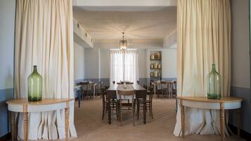 A dining area with a long table and chairs, surrounded by cream-colored curtains, and two small tables with green bottles.