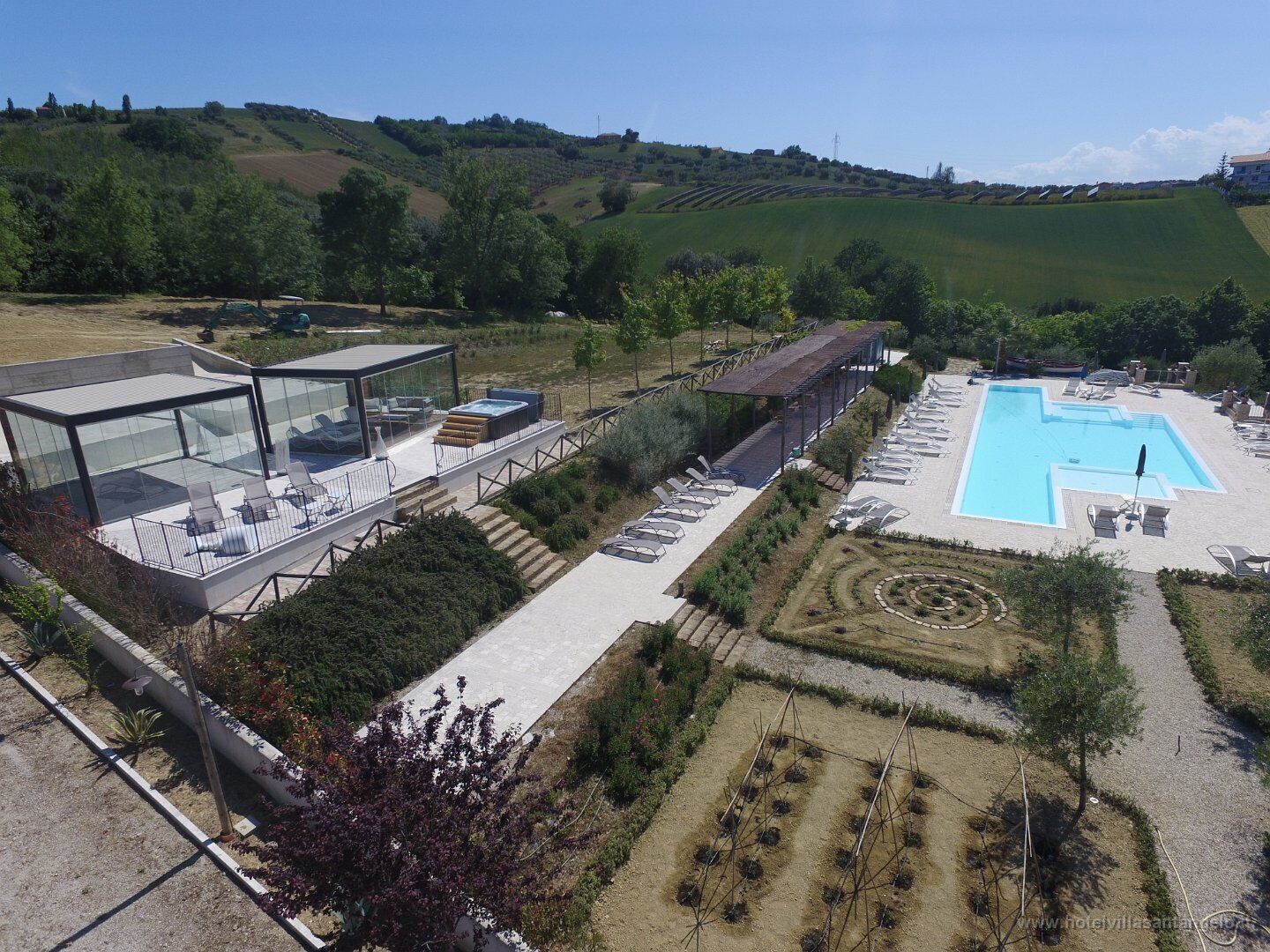 A modern hotel with a glass-enclosed pool area, outdoor seating, and a view of the surrounding countryside.