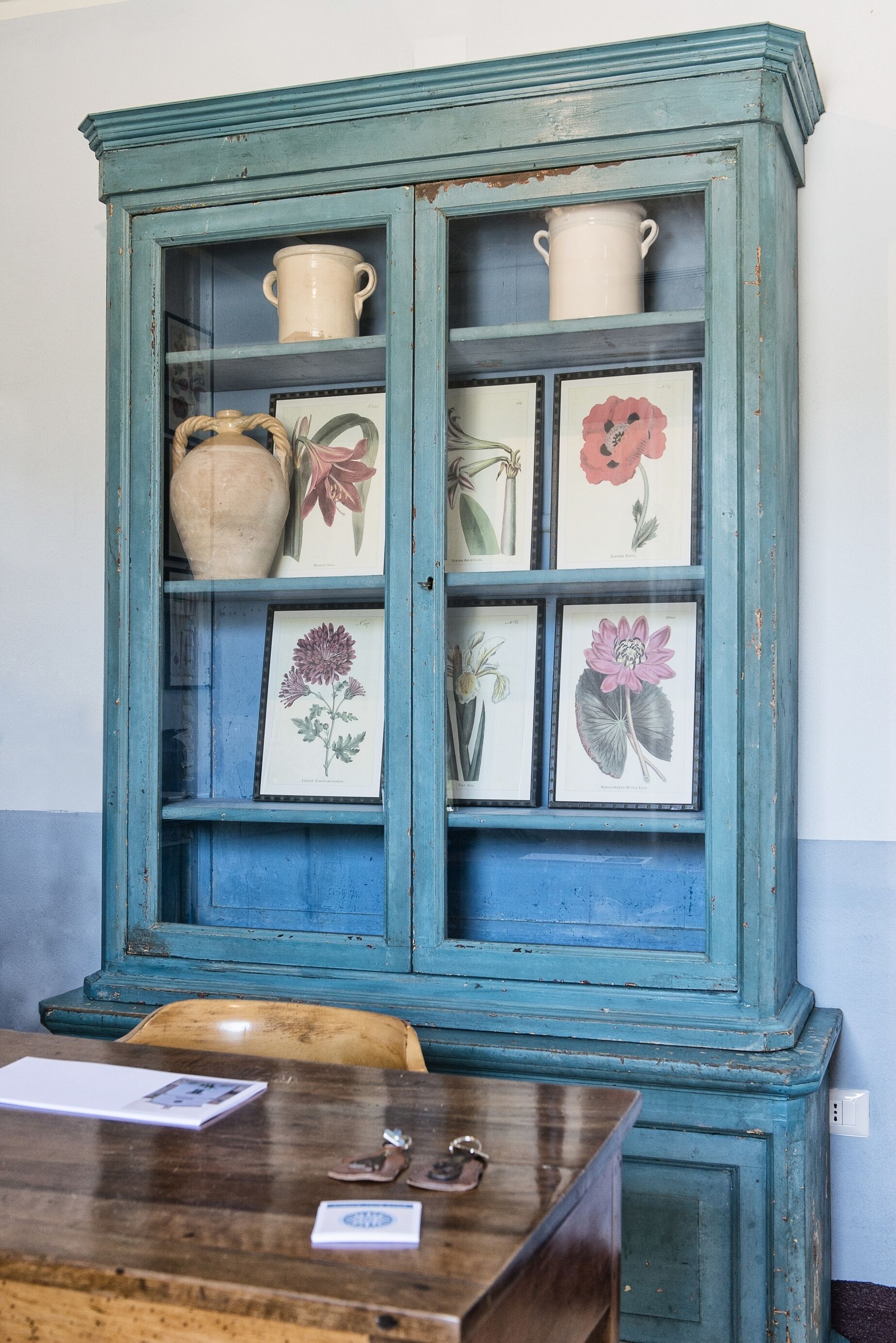 A teal cabinet with glass doors, displaying various ceramic items and framed botanical illustrations.