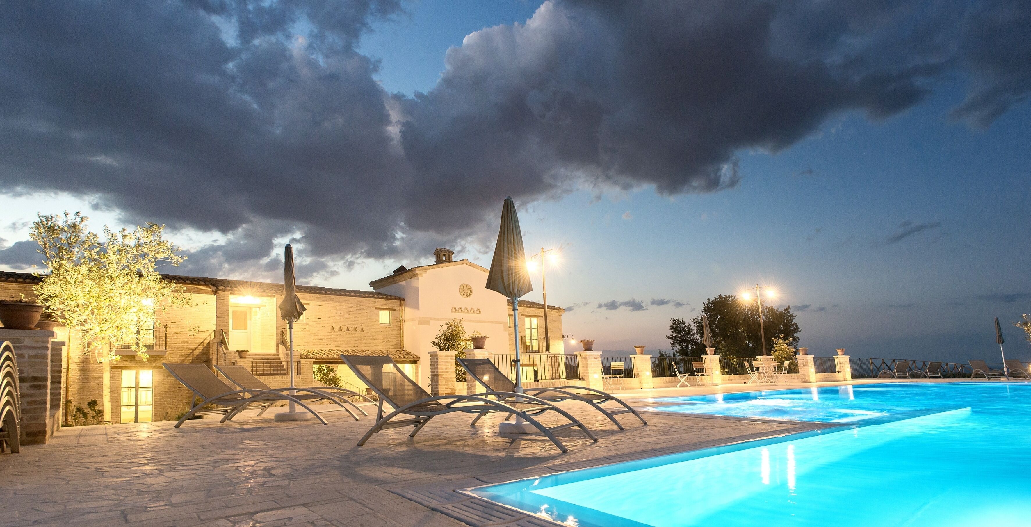 Une piscine avec des chaises longues, un bâtiment doté d’une horloge et un ciel nuageux.