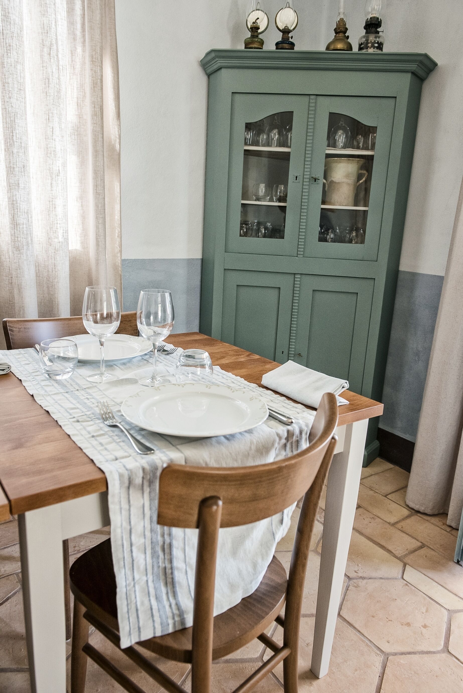 Un tavolo da pranzo apparecchiato con piatti, bicchieri e posate, una sedia in legno e un mobile contenitore verde con vetrine.