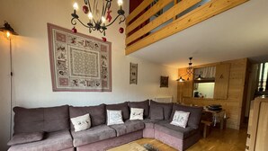Un soggiorno accogliente con un divano componibile, un tavolino in legno e un tappeto decorato. Si intravede una cucina sullo sfondo, con pensili in legno e un lampadario.