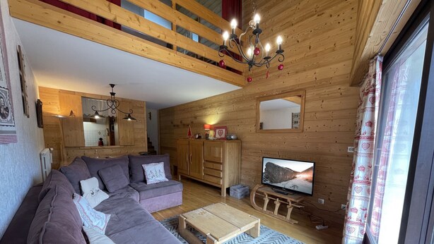 Un accogliente soggiorno con un divano componibile, travi a vista in legno, un lampadario, una televisione e un'ampia finestra con tende.