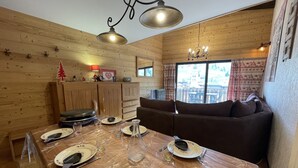 Un accogliente interno in legno con un tavolo da pranzo per due, un divano e vista su un paesaggio montuoso attraverso ampie finestre.