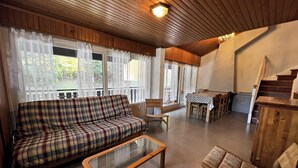 A cozy living room with a plaid sofa, a glass-top coffee table, wooden chairs, a dining table with a polka dot tablecloth, and a staircase. - BELMONT 26 – Village-center duplex close to the slopes (La Clusaz)