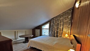 A bedroom with a wooden floor, a bed with a white bedspread, a nightstand with a lamp, a window with curtains, and a radiator. - BELMONT 26 – Village-center duplex close to the slopes (La Clusaz)