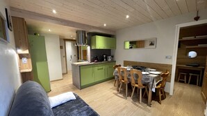 A modern living room with a grey sofa, wooden dining table, and chairs. A kitchen with green cabinets and a stove is visible in the background. - GENTIANES 3 – Apartment in the village center and close to ski slopes – Wi-Fi (La Clusaz)