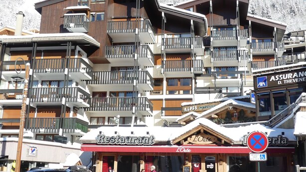 Een meerhoog gebouw met balkons, een restaurant genaamd "Restaurant L'Otta", en een bord met "Alpes Immobilier".