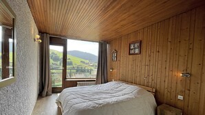 A bedroom with a large bed, wooden walls, a window with a view of a green landscape, and a small stool.