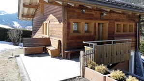 A wooden cabin with a sloped roof, wooden steps leading to a porch, and a view of snow-capped mountains in the background. - PETIT LUTIN – Chalet on the slopes and in the village center – Garage and parking (La Clusaz)