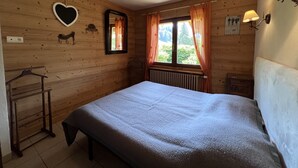 A wooden-paneled room with a bed, a radiator, a window with curtains, a wall-mounted lamp, and a coat rack. - PETIT LUTIN – Chalet on the slopes and in the village center – Garage and parking (La Clusaz)