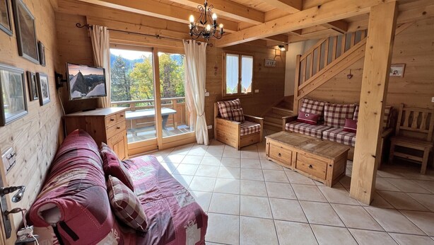 Ein Interieur einer Holzhütte mit einer Couch, einem Fernseher, einer Treppe und Blick ins Freie.