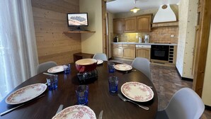 Zona pranzo con tavolo in legno apparecchiato per quattro persone, televisione a parete e cucina visibile tramite un'apertura.