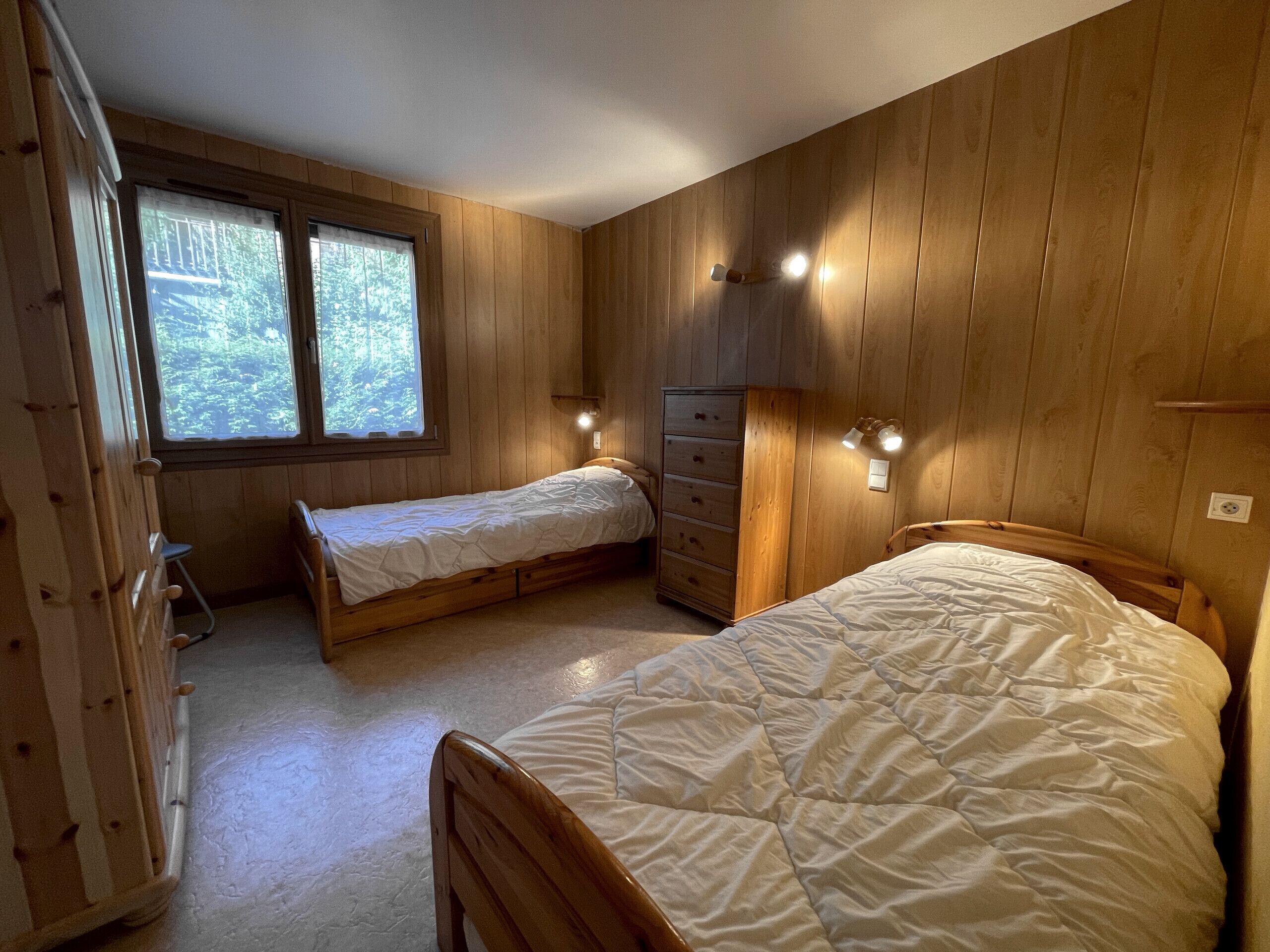 Una habitación con dos camas de madera, un aparador y una ventana con vistas a árboles.