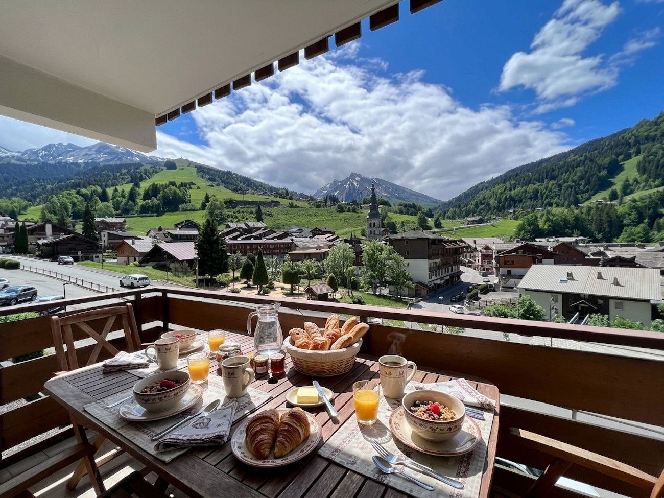 A balcony with a table set for breakfast, overlooking a village and mountains.