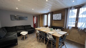 Un salon moderne avec un canapé d’angle noir, une table à manger en bois pour quatre personnes et un grand téléviseur fixé au mur.