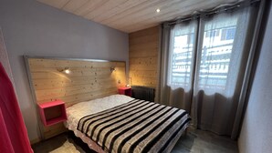 Une chambre à coucher avec une tête de lit en bois, un lit recouvert d’une couverture à rayures, une table de chevet rouge, une petite table et une fenêtre avec des rideaux.