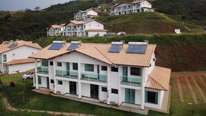 A modern residential building with multiple floors, solar panels on the roof, and a hillside backdrop. - Casa no Mirante de Escarpas (Capitólio)