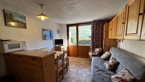 A compact kitchen with a microwave, wooden cabinets, a dining table, and a sofa with patterned cushions. - BIBLOS 15 - Apartment in the village center, close to the slopes - Parking (La Clusaz)