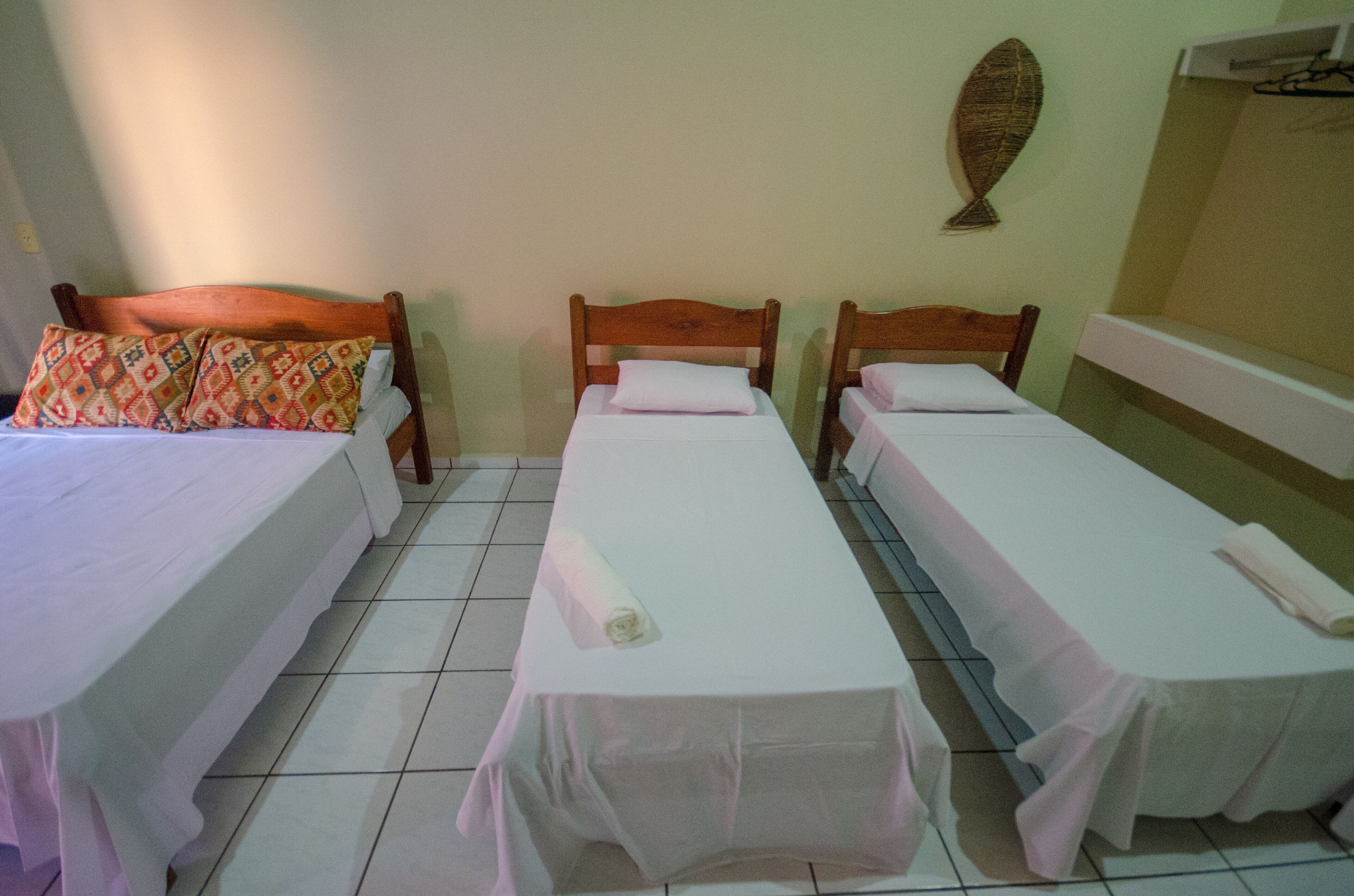 Una habitación con tres camas individuales, cada una con una colcha y almohadas blancas. El suelo es de baldosas y hay un estante fijado a la pared con un objeto decorativo.