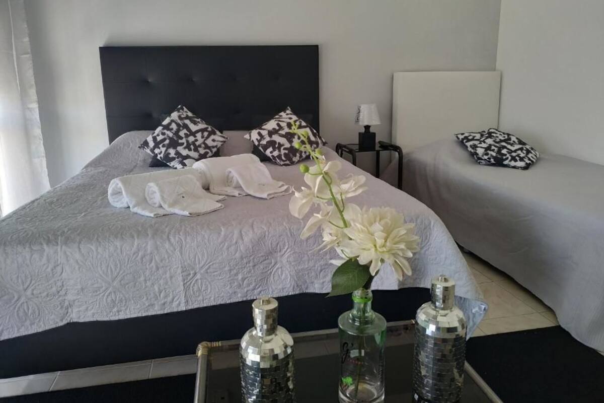 Ein ordentlich bezogenes Bett mit weißer Decke, zwei gemusterten Kissen und einer Vase mit weißen Blumen. Zwei Nachttische sind vorhanden, einer mit einer Lampe und der andere mit einer Flasche.