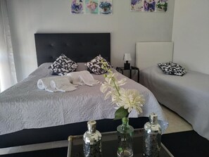 A neatly made bed with a white comforter, two patterned pillows, and a vase with white flowers. There are two bedside tables, one with a lamp and the other with a bottle.