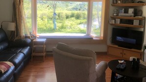 Ein Wohnzimmer mit großem Fenster, Deckenventilator, schwarzem Ledersofa, beigem Sessel, Fernseher und einem kleinen Tisch mit Fernbedienungen und Flaschen.