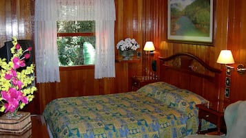 A wooden room with a bed, a window with lace curtains, a painting on the wall, and a nightstand with a lamp.