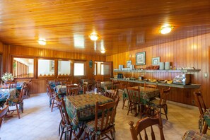 Uma sala de jantar com painéis de madeira, mesas e cadeiras, bancada de cozinha com eletrodomésticos e vista pela janela.
