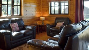 L’intérieur d’un chalet confortable, avec des canapés en cuir, une petite table avec une lampe et de grandes fenêtres ornées de rideaux.