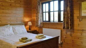 Une pièce de cabane en bois comprenant un lit, une table de chevet, une lampe et une fenêtre avec des rideaux.