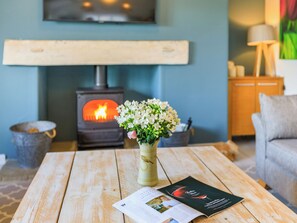 Un salón acogedor con una mesa de madera, un jarrón de flores, una revista y una estufa de leña al fondo.