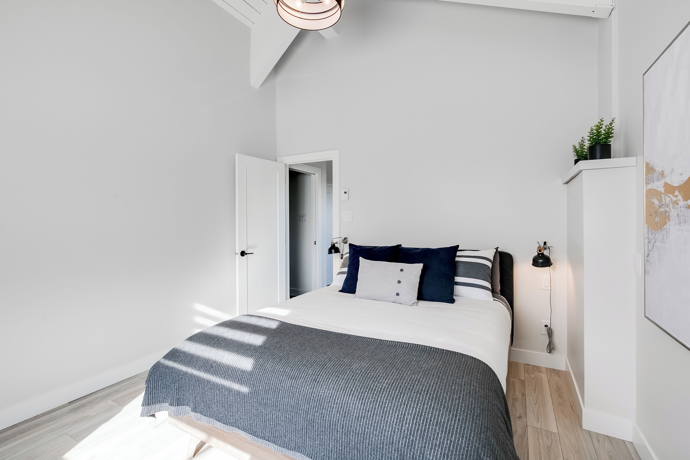 A modern bedroom with a large bed, a bedside table with a lamp, and a door leading to another room.