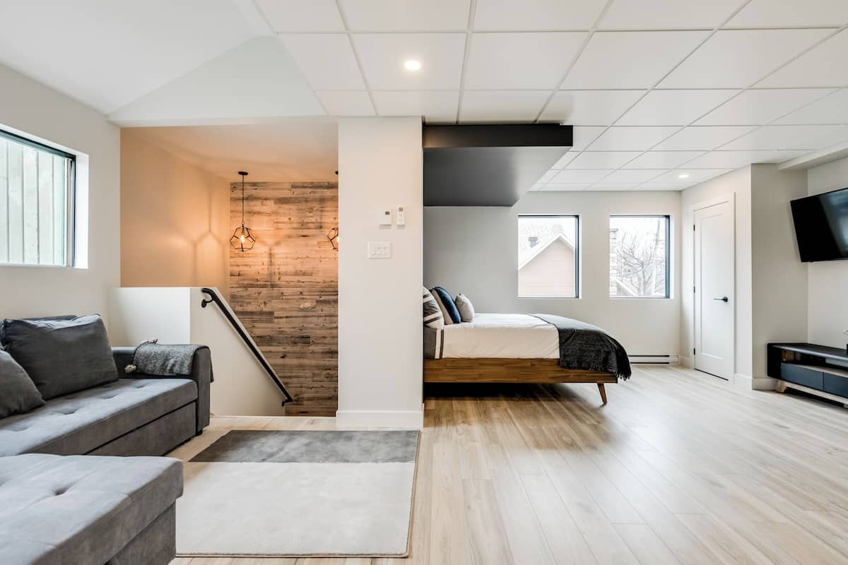 A modern living room with a grey sofa, a wooden staircase, and a bed with a white headboard.