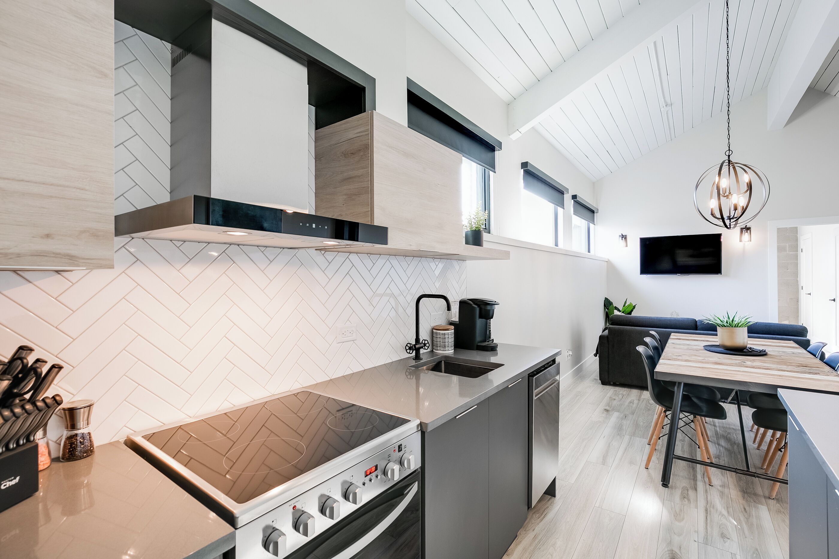 Een moderne keuken met een inbouwfornuis, spoelbak en vaatwasser. Er is een eethoek met een tafel en stoelen, en een woonkamer met een bank en een televisie.
