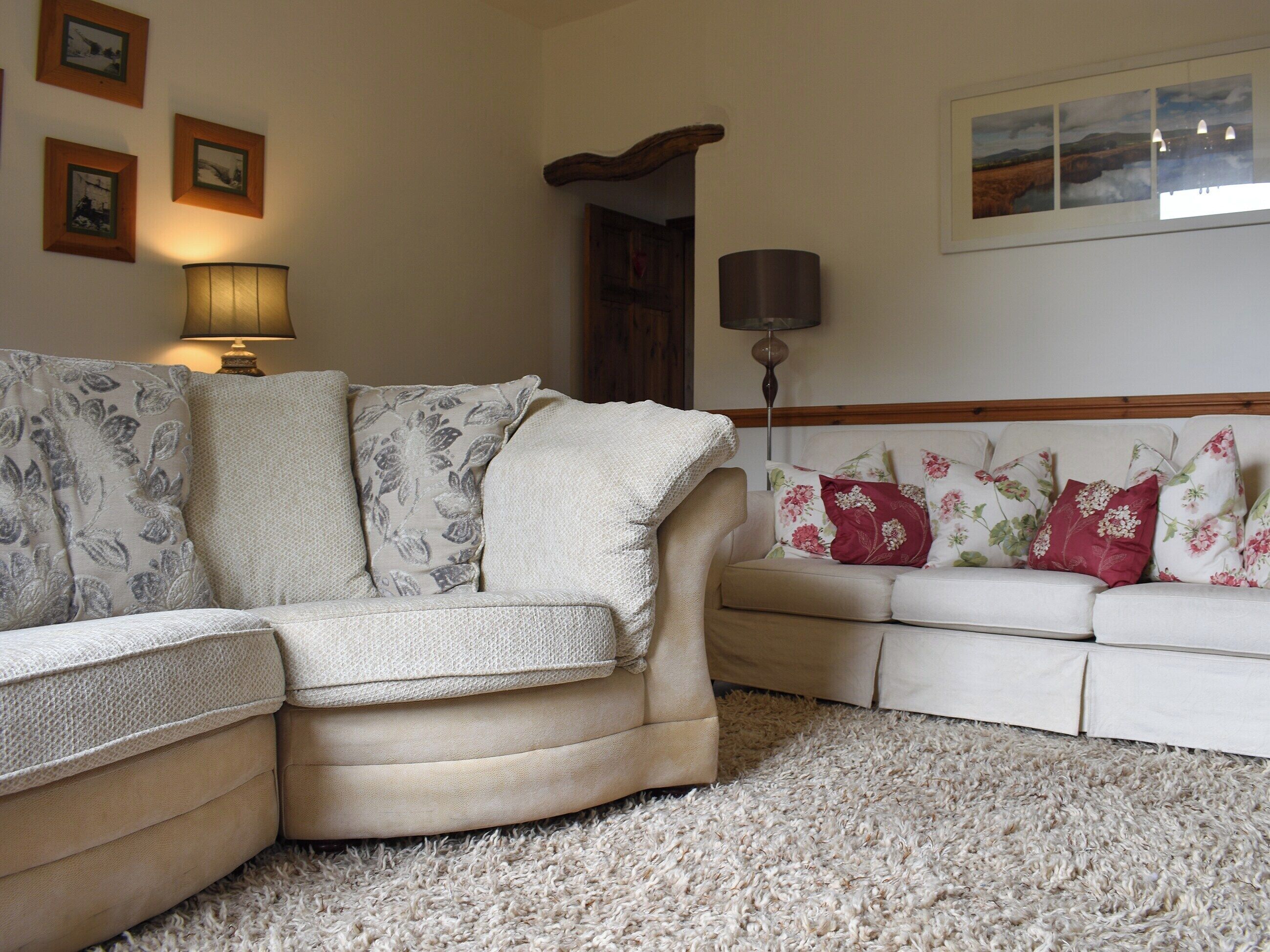 A living room with a beige sectional sofa, a white sofa with floral cushions, a shaggy carpet, and wall-mounted lamps.
