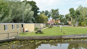 A lakeside view with a row of mobile homes, a picnic table, and a calm water surface with lily pads.