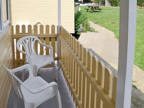 A balcony with two white plastic chairs, a yellow picket fence, and a view of a picnic table and grass.