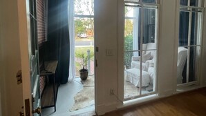 Una habitación con suelo de madera, puerta corredera de vidrio que da a un espacio exterior con una silla blanca y una planta en maceta.
