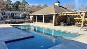 Une piscine rectangulaire entourée d’une terrasse en pierre, avec un patio couvert et un bâtiment doté de grandes fenêtres en arrière-plan.