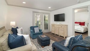 Un salon comprenant un canapé, un fauteuil, un pouf, une télévision, et une chambre adjacente avec un lit.