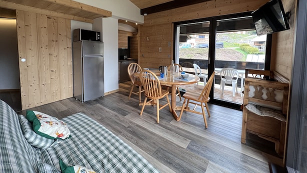 A cozy cabin interior with a wooden dining table, chairs, a sofa, a kitchen with a refrigerator and microwave, and a TV mounted on the wall. - SAPAUDIA G – Ski-in/ski-out apartment, lovely view – Parking (La Clusaz)