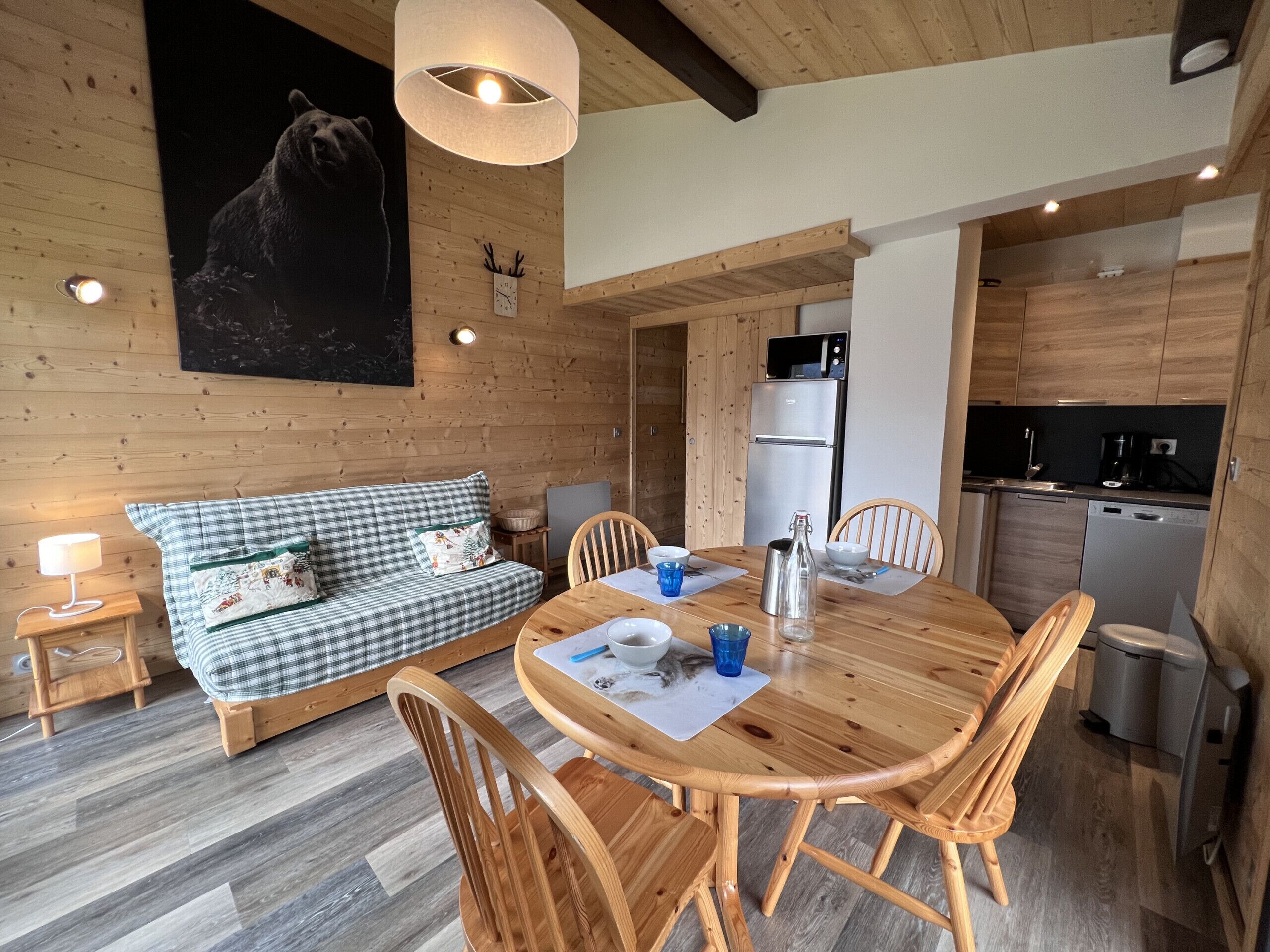 A cozy cabin interior with a wooden dining table, chairs, a sofa, a kitchen area, and a bear mural on the wall.